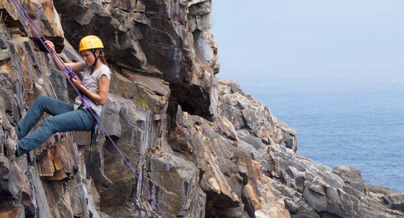 A person wearing safety gear is secured by ropes as they rock climb. They are high above a blue body of water. 